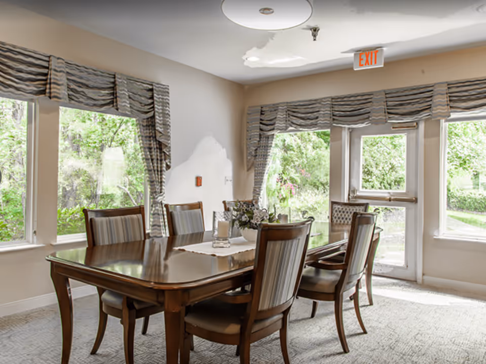 A bright dining room with a wooden dining table and six chairs with striped upholstery. The room has large windows with patterned valances, allowing natural light to fill the space and providing a view of green trees outside. There is an exit door with a red EXIT sign above it.