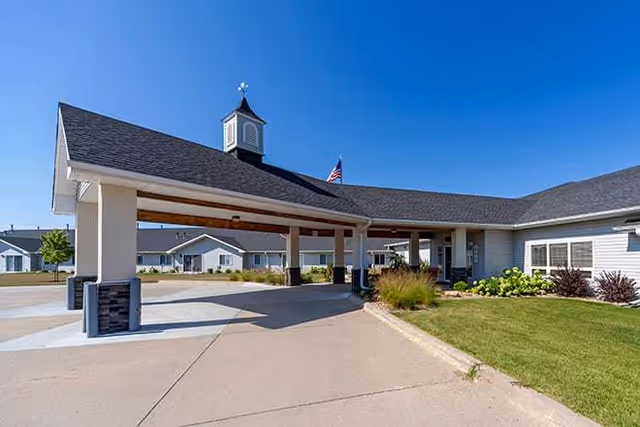 Entrance area of a senior living facility with a covered drop-off zone, manicured lawn, and clear blue sky. The building has a gray roof with a small cupola and an American flag flying near the entrance.