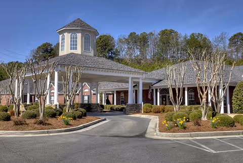 Exterior view of a single-story brick building with a covered entrance and a small tower-like structure on the roof, surrounded by landscaped bushes and trees under a clear blue sky.