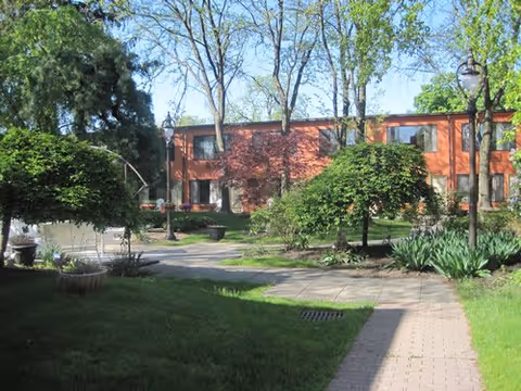 Sunlit landscaped courtyard with paved walkways, benches, trees and shrubs in front of a two-story brick residential building.