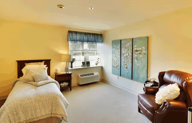 A cozy bedroom with a single bed covered in light-colored bedding, a wooden nightstand with a lamp and decorative items, a window with a blue valance, a wall-mounted air conditioning unit below the window, a three-panel floral artwork on the wall, and a brown leather armchair with a white fluffy pillow.