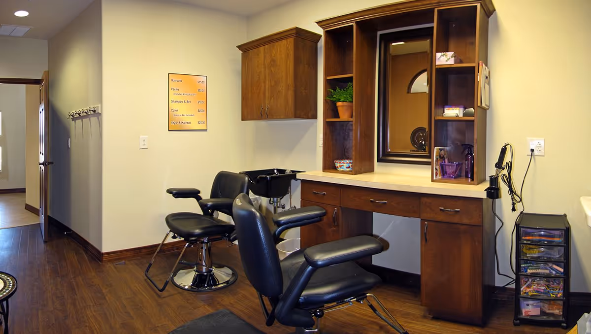 Interior salon area with two styling chairs, wooden cabinetry and a mirror in an assisted living facility.
