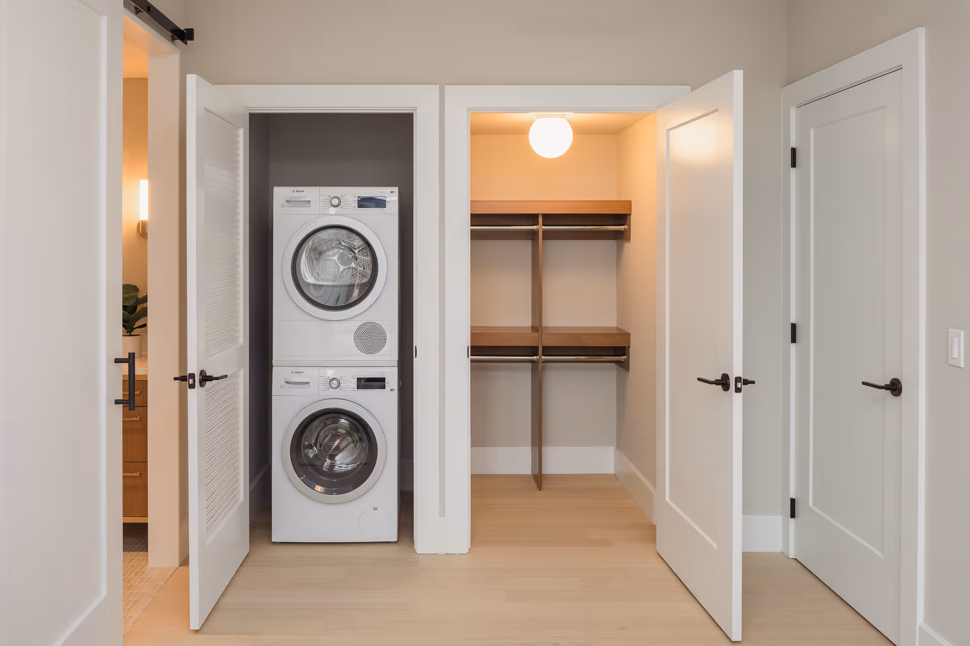 A modern interior space featuring an open closet with a stacked washer and dryer on the left and an empty walk-in closet with wooden shelves and hanging rods on the right. The walls are painted light beige, and the floor is light wood. There are three white doors with black handles, one partially open showing a glimpse of a bathroom with a wooden vanity and a plant.