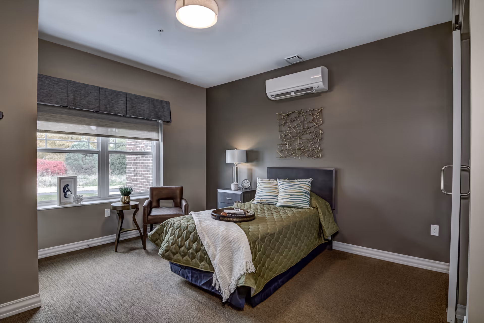 A cozy bedroom with a single bed covered in a green quilt and two striped pillows. A white throw blanket is draped over the bed with a tray holding a glass and books. Next to the bed is a nightstand with a lamp and a clock. A brown leather chair and small round table with a plant are positioned near a window with gray blinds. The walls are painted taupe, and a decorative metal wall art hangs above the bed. A white air conditioning unit is mounted on the wall above the bed.