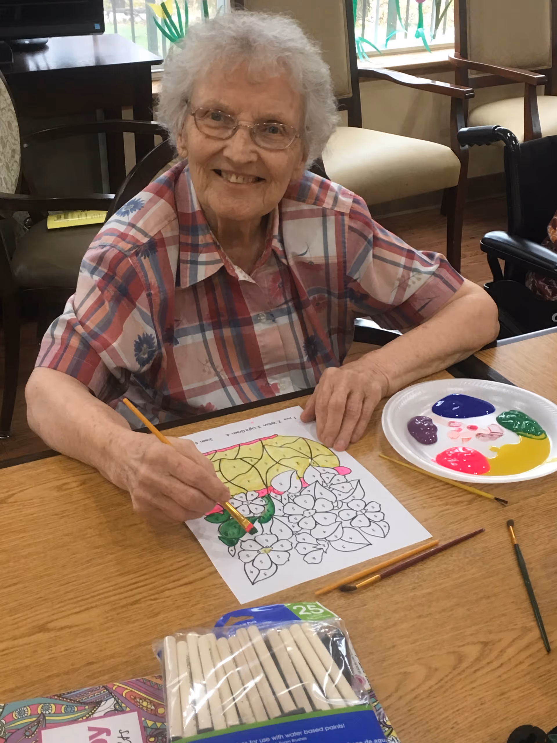 An elderly woman with glasses and a plaid shirt is sitting at a wooden table, smiling while painting a floral coloring page with green paint. On the table are paintbrushes, a palette with various paint colors, and a pack of white chalk pastels. The background shows chairs and windows with natural light.