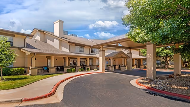 Exterior view of Brookdale Remington Park senior living facility showing a large building with a covered entrance driveway, surrounded by trees and landscaping under a partly cloudy sky.