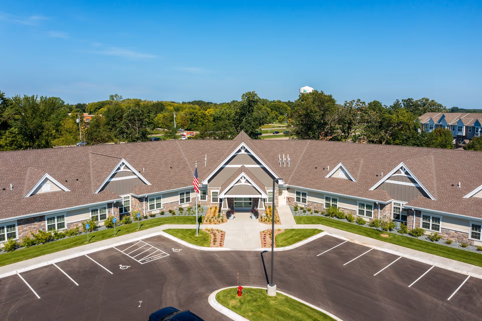 Aerial view of NorBella Senior Living - Champlin building with a large parking lot in front, an American flag on a flagpole, and landscaped greenery surrounding the entrance. The building has a brown roof, stone and beige siding, and multiple windows under a clear blue sky.