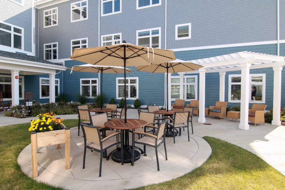 Outdoor patio area at Benchmark Senior Living on Clapboardtree featuring round wooden tables with beige umbrellas and matching chairs. There is a white pergola with wicker chairs underneath, surrounded by a well-maintained garden and the exterior of a blue and white building with multiple windows.