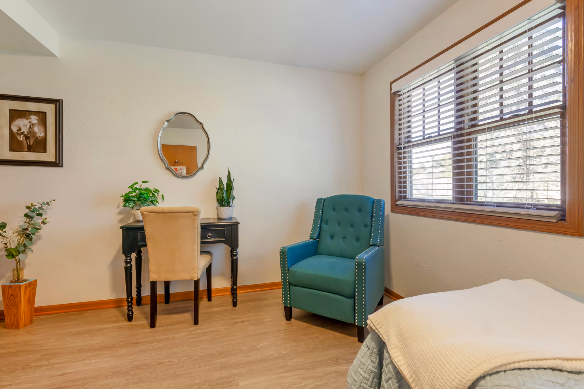 Bedroom corner with a teal armchair, small black desk and beige chair, potted plants, a bed corner, and a window with blinds.