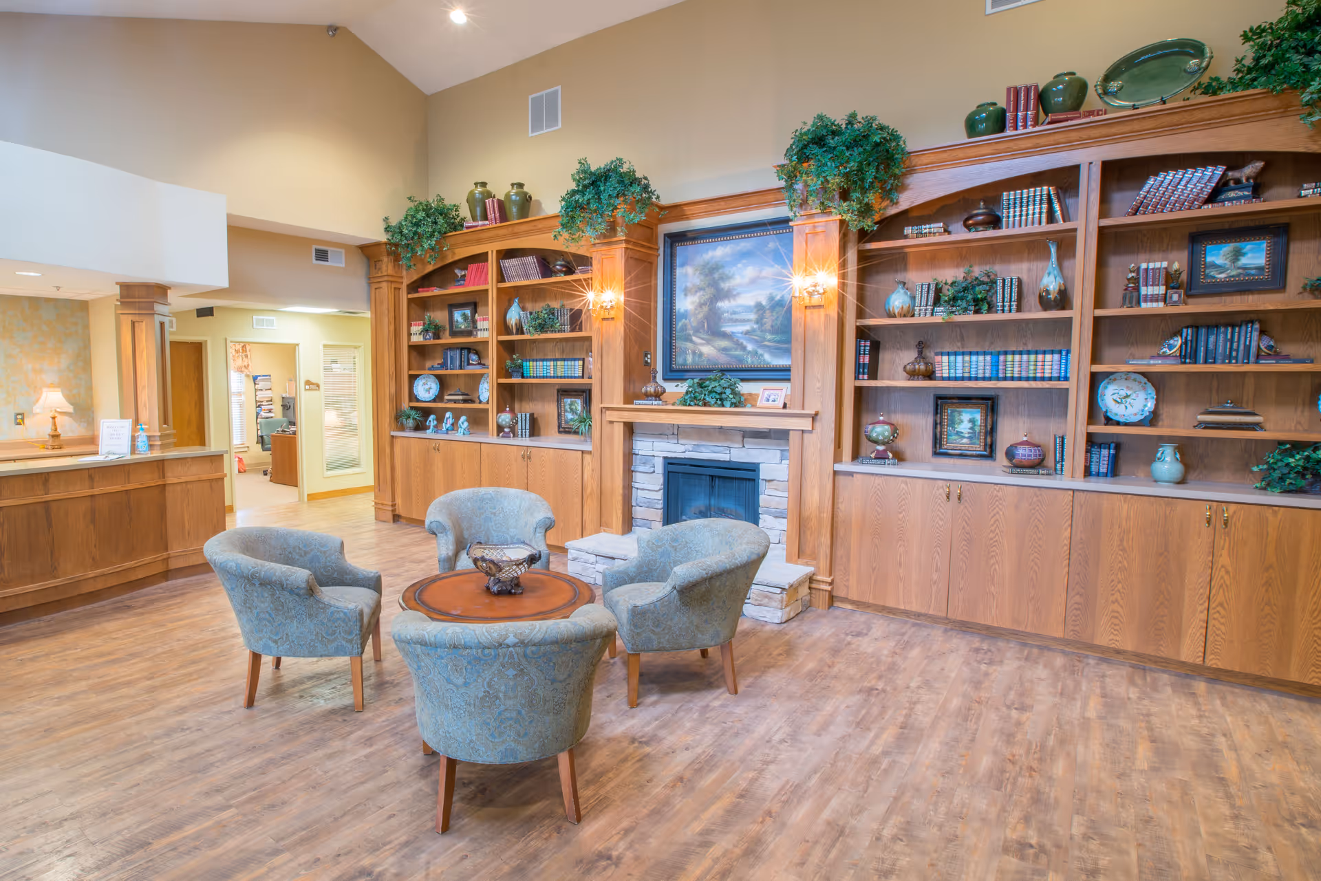 A cozy common area in a nursing center featuring four upholstered armchairs arranged around a round wooden table. Behind the seating area is a stone fireplace with a large framed painting above it. Flanking the fireplace are built-in wooden bookshelves filled with books, decorative plates, vases, and plants. The room has warm wooden flooring and soft lighting, creating a welcoming atmosphere.