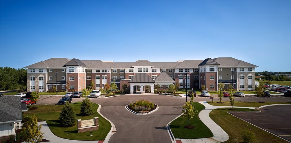 Front exterior view of Alden Estates Cts Of Huntley, a large three-story senior living facility with a circular driveway, landscaped greenery, and parked cars under a clear blue sky.