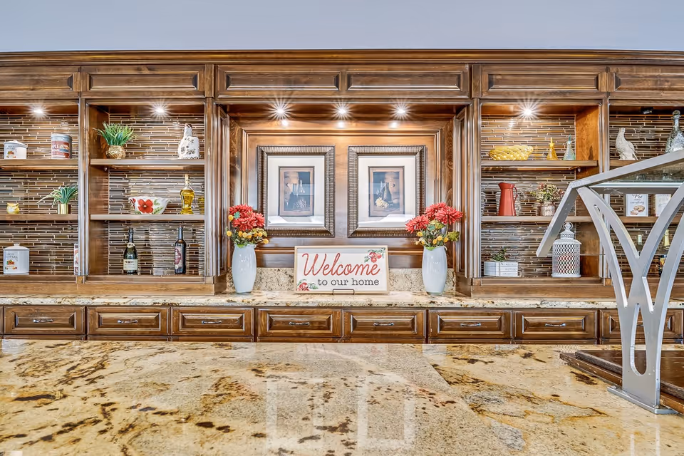A wooden cabinet with shelves displaying decorative items including plants, bottles, and containers. In the center, there are two framed pictures above a countertop with two white vases holding red flowers and a sign that says 'Welcome to our home'. The countertop is made of polished stone with a beige and brown pattern.