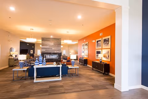A spacious and well-lit living room area with modern decor. The room features a central fireplace with a stone surround, flanked by sofas and tables with decorative lamps and vases. The far wall is painted orange and adorned with framed pictures. The flooring is carpeted with a patterned design, and the ceiling has recessed lighting and hanging light fixtures.