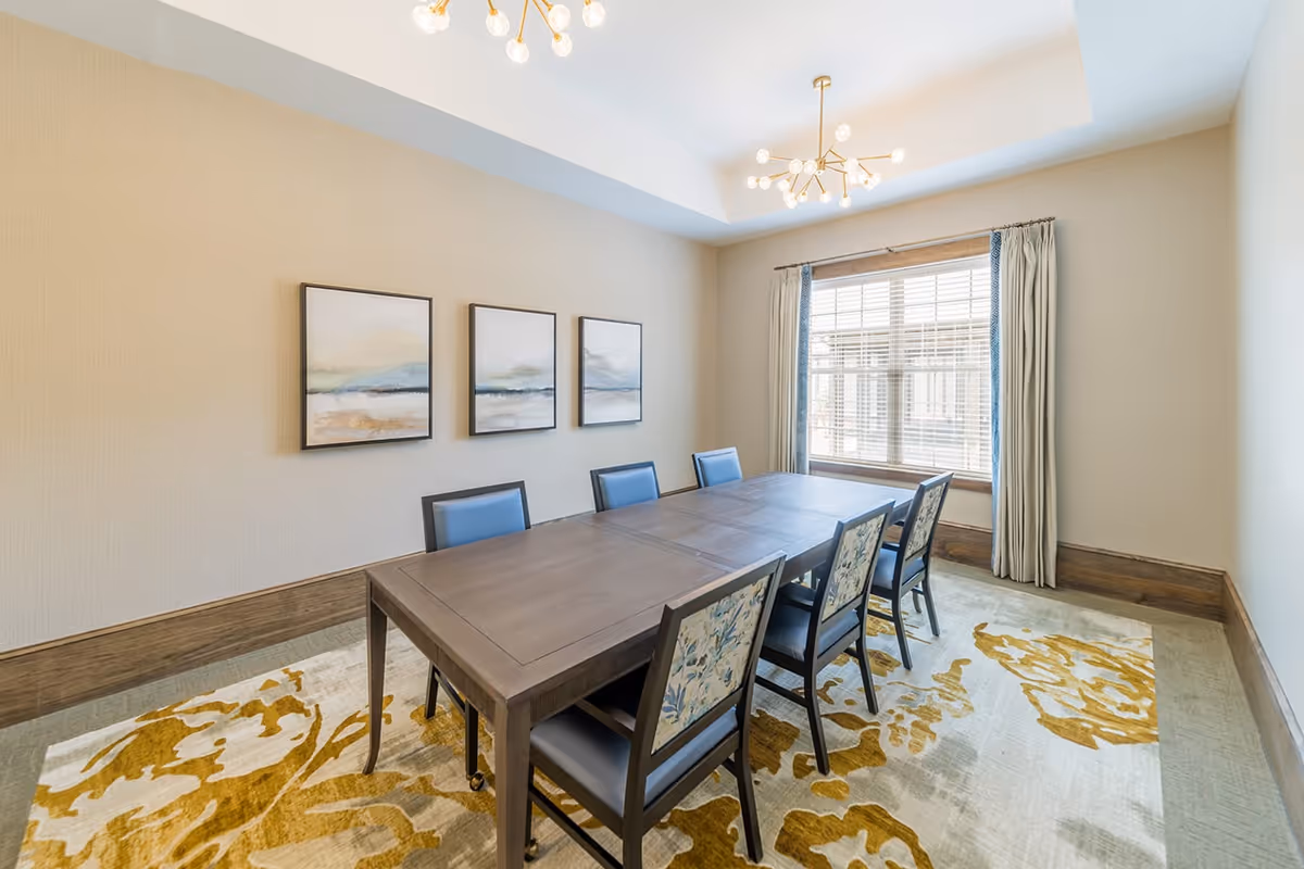 A bright dining room with a large wooden table surrounded by six chairs, some with blue cushions and others with floral patterned backs. The room features a large window with white blinds and curtains, three framed abstract landscape paintings on the wall, a beige textured wallpaper, and a decorative yellow and white patterned rug. Two modern chandeliers with multiple light bulbs hang from the ceiling.