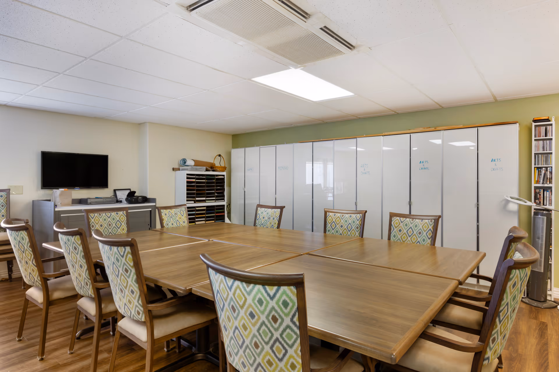 A senior facility activity room with a large table surrounded by patterned chairs, storage cabinets, and a wall-mounted TV.