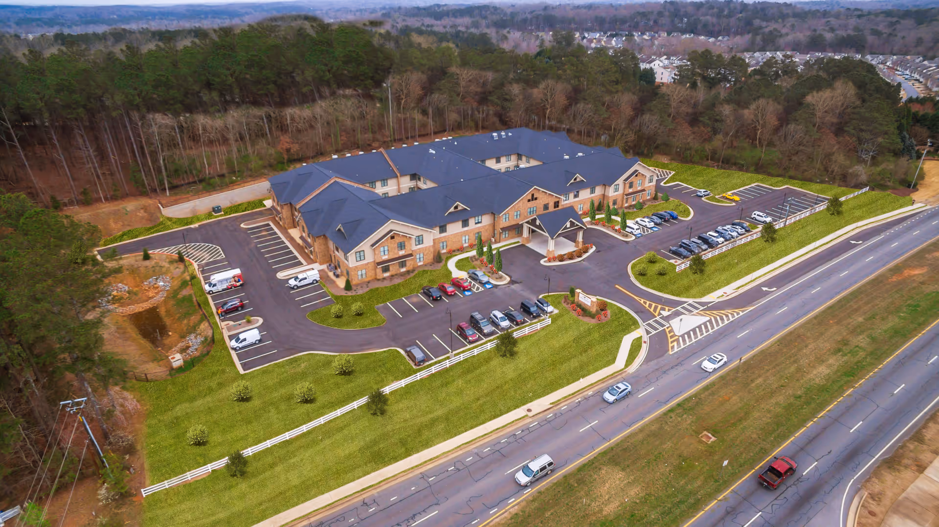 Aerial view of a large senior living facility named Brickmont at Woodstock, showing a two-story building with a dark roof surrounded by parking lots, green lawns, and trees. The facility is adjacent to a multi-lane road with several cars driving by, and there is a wooded area behind the building.
