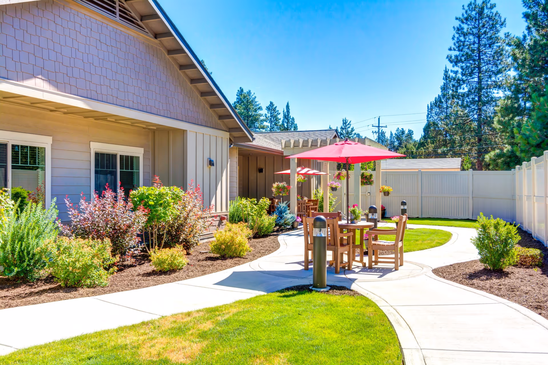 Sunny courtyard with paved walkways, patio tables and red umbrellas, landscaped beds, and a single-story building facade.