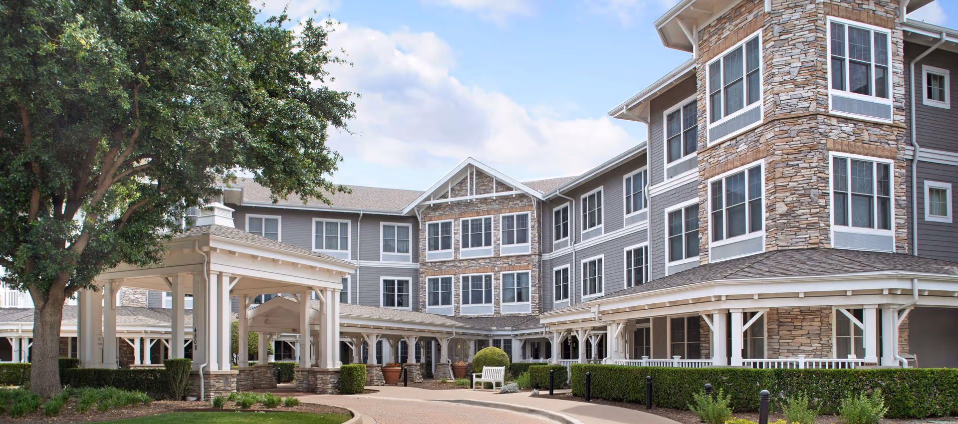 Exterior view of a senior living facility building with stone and gray siding, multiple windows, a covered entrance, and landscaped greenery including a large tree and bushes.