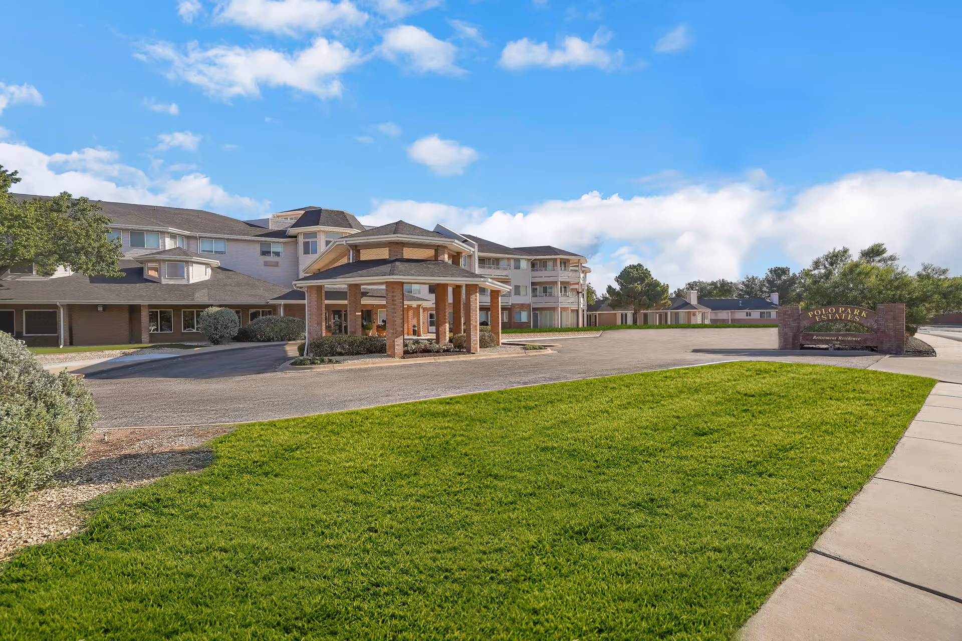 Exterior view of Polo Park Estates senior living facility showing a large building with multiple floors, a covered entrance with brick pillars, a well-maintained lawn, and a sign with the facility name near the driveway under a blue sky with some clouds.