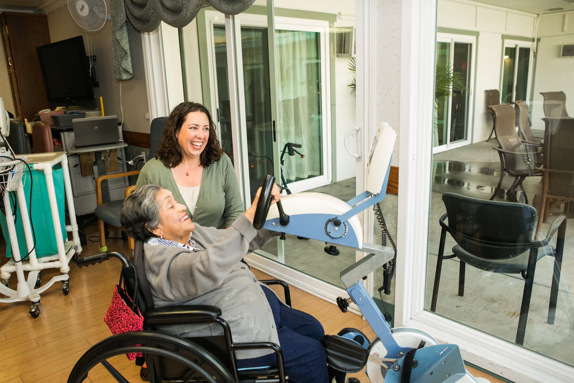 An elderly woman in a wheelchair uses a hand cycle exercise machine indoors near a large glass sliding door, while a smiling caregiver watches and supports her.