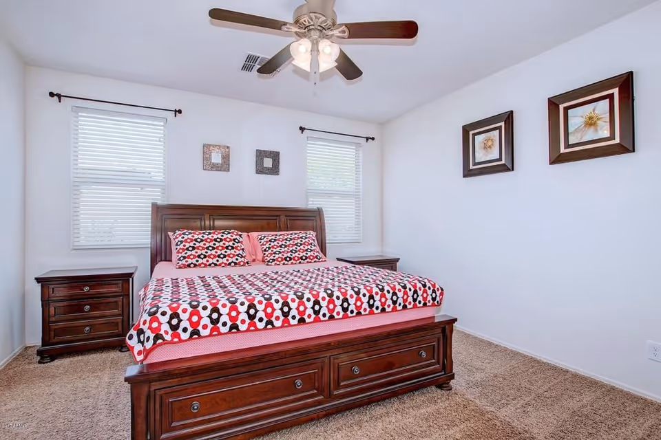 Bright bedroom with dark wood bed, two nightstands, three windows, and ceiling fan.