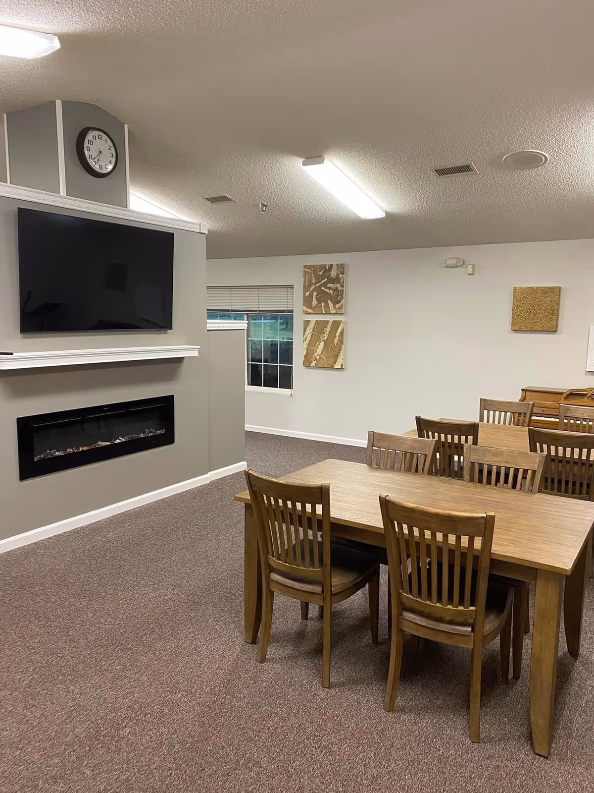 Interior room with wooden tables and chairs arranged in rows, a wall-mounted flat screen TV above a modern electric fireplace, a clock on the wall, and abstract artwork hanging on the walls.