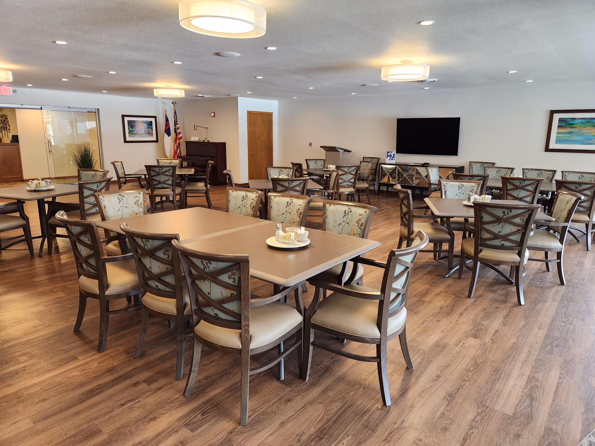 Spacious communal dining room with multiple tables and chairs, wood flooring, and a wall-mounted TV.