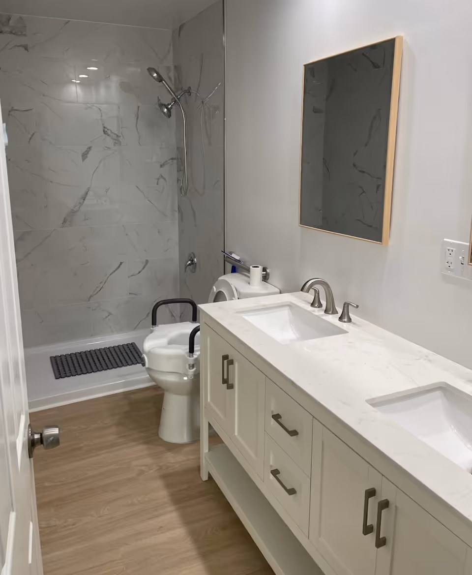 A modern senior-accessible bathroom with a double-sink vanity, wall mirror, toilet with safety rails, and a tiled walk-in shower.