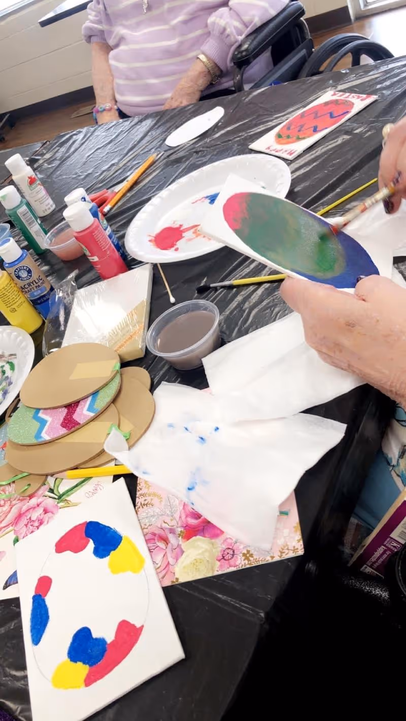 Two elderly individuals seated at a table covered with a black plastic tablecloth, engaged in painting colorful Easter egg crafts using acrylic paints and brushes. Various art supplies including paint bottles, paper plates with paint, and decorated cardboard egg shapes are spread across the table.