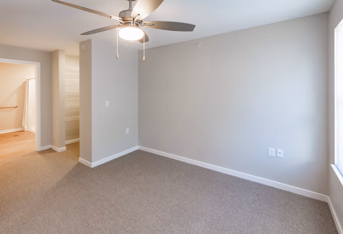 Empty carpeted bedroom with a ceiling fan, neutral walls, and an open doorway to a bathroom/closet.