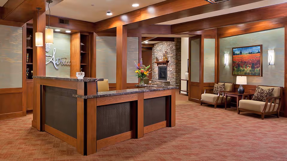 Reception area of Atria Valley View featuring a wooden front desk with a marble countertop, a vase with flowers, comfortable seating with armchairs and side tables, a stone fireplace in the background, warm lighting, and a framed painting on the wall.