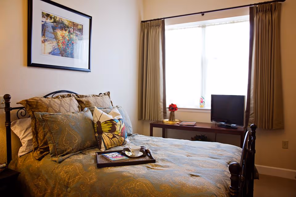 A cozy bedroom with a neatly made bed featuring multiple pillows, including one with a butterfly design. A tray with a cup and a book rests on the bed. There is a framed picture on the wall above the bed and a window with curtains letting in natural light. A small table beneath the window holds a vase with red flowers and a flat-screen TV.