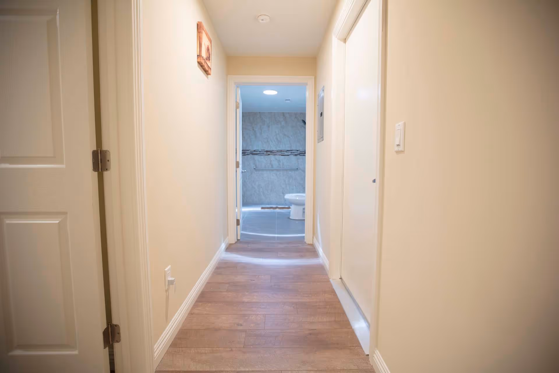 View down a hallway with light-colored walls and wooden flooring leading to a bathroom with a visible toilet and tiled walls. There is a small framed picture on the left wall and a closed door on the right side of the hallway.