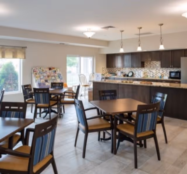 A bright dining area in a senior living facility with several wooden tables and chairs arranged neatly. In the background, there is a kitchen area with dark wood cabinets, a tiled backsplash, and a countertop with pendant lights hanging above. Large windows allow natural light to fill the room.