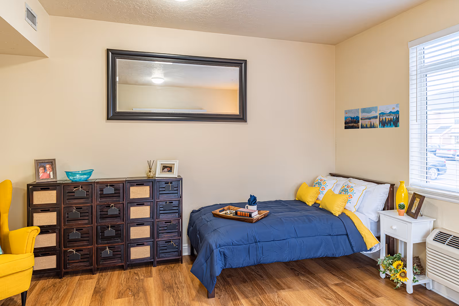 Furnished bedroom with a blue bed and yellow pillows, a multi-drawer dresser, wall mirror, and a window.