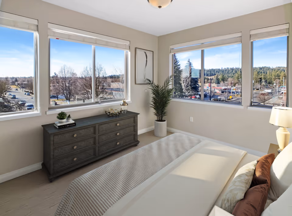 A bright bedroom with large windows showing an outdoor view of trees and buildings. The room features a bed with beige and brown pillows, a dark wooden dresser with decorative items, a potted plant in the corner, and a lamp on a bedside table.