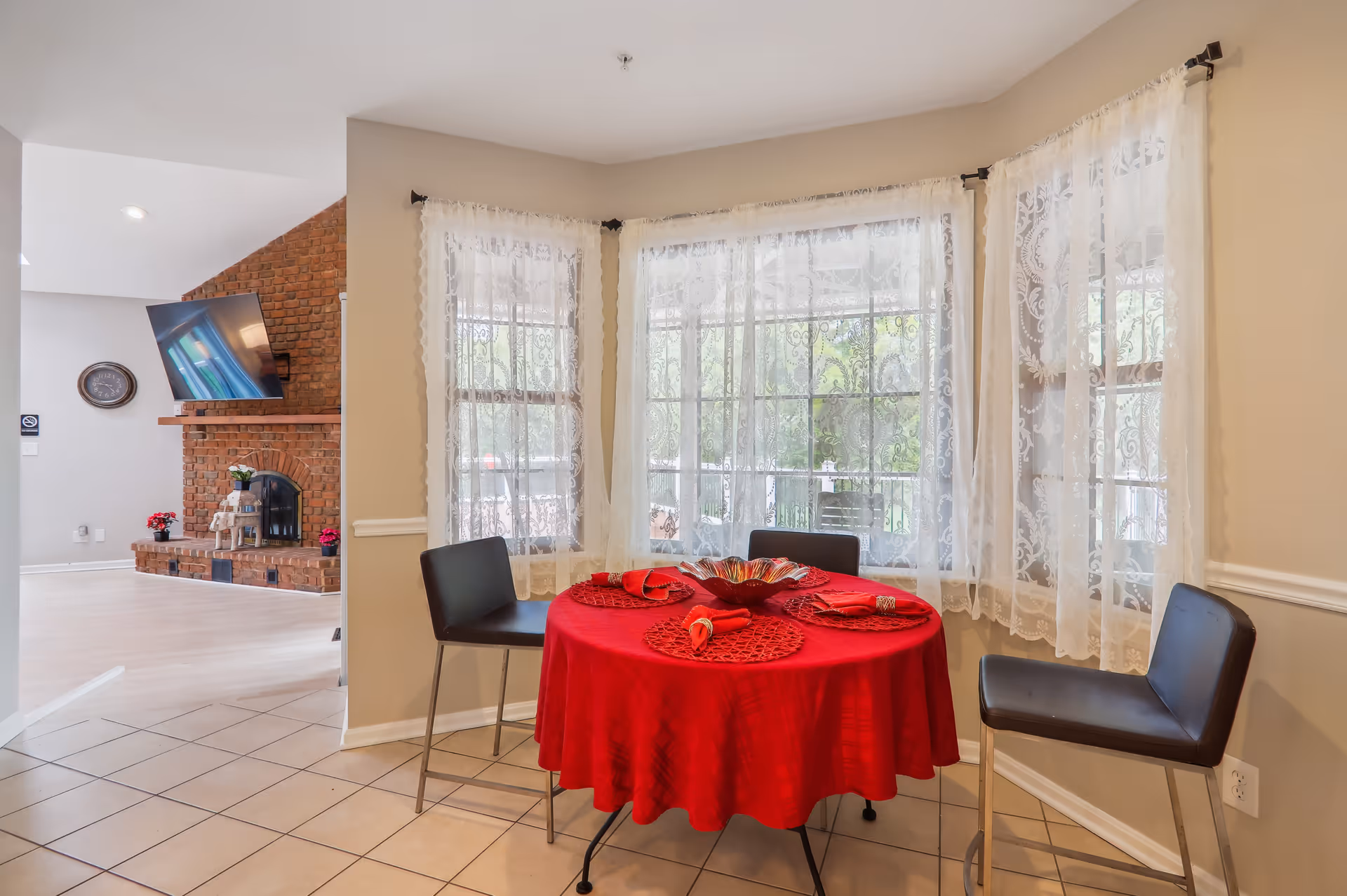 A small dining area with a round table covered with a red tablecloth and set with red placemats and napkins. Three black chairs surround the table. The area is next to large windows with sheer white lace curtains, allowing natural light to fill the space. In the background, there is a living room with a brick fireplace, a mounted TV, and some decorative plants.