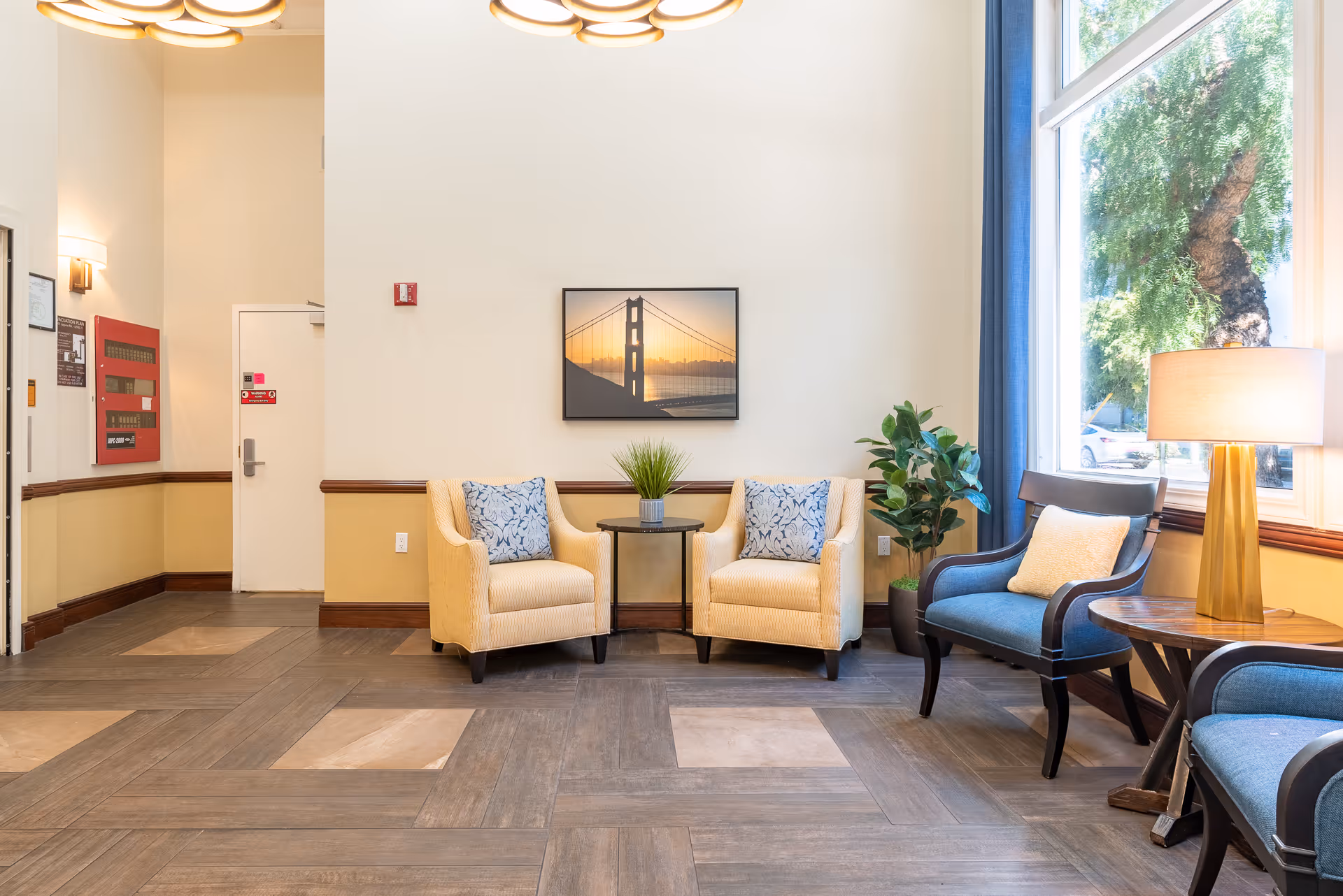 A bright and inviting seating area in a senior living facility featuring two beige armchairs with blue patterned pillows, a small round table with a plant between them, and two blue cushioned chairs with a wooden side table and lamp near a large window with blue curtains. A framed picture of the Golden Gate Bridge hangs on the wall above the armchairs.