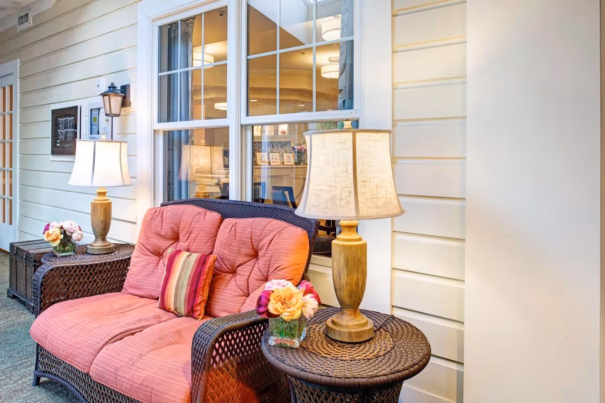 A cozy seating area with a cushioned wicker loveseat in a soft coral color, accompanied by a striped throw pillow. On either side of the loveseat are round wicker side tables, each holding a table lamp with a beige shade and a small vase of flowers. The setting is indoors, next to a window with white framing, and the walls are painted in a light cream color with horizontal paneling.