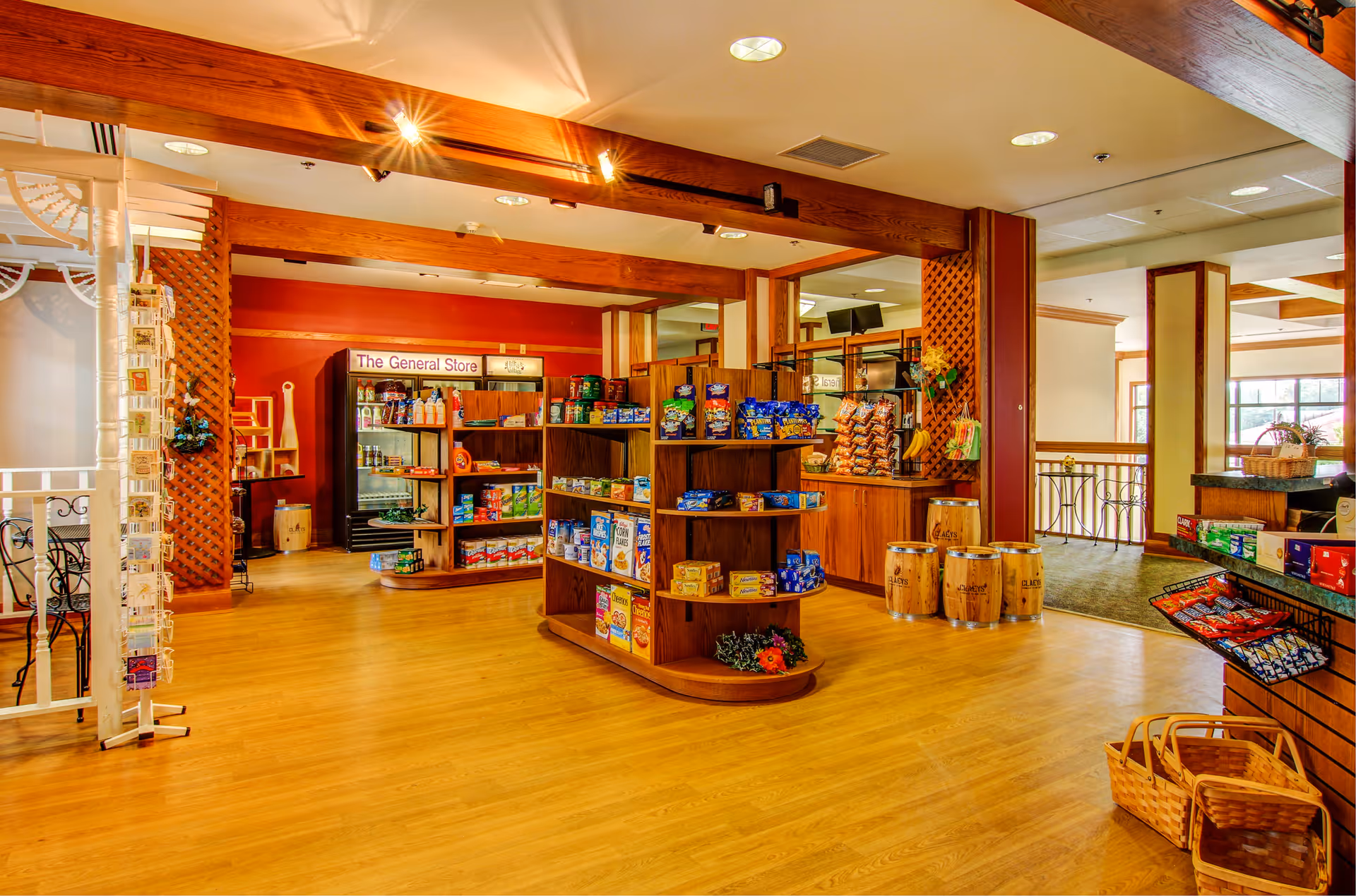 Interior view of a general store area inside a senior living facility with wooden shelves stocked with snacks, beverages, and other grocery items. The space features wooden beams, a light wood floor, and warm lighting. There is a sign that reads 'The General Store' above a refrigerated section in the background.