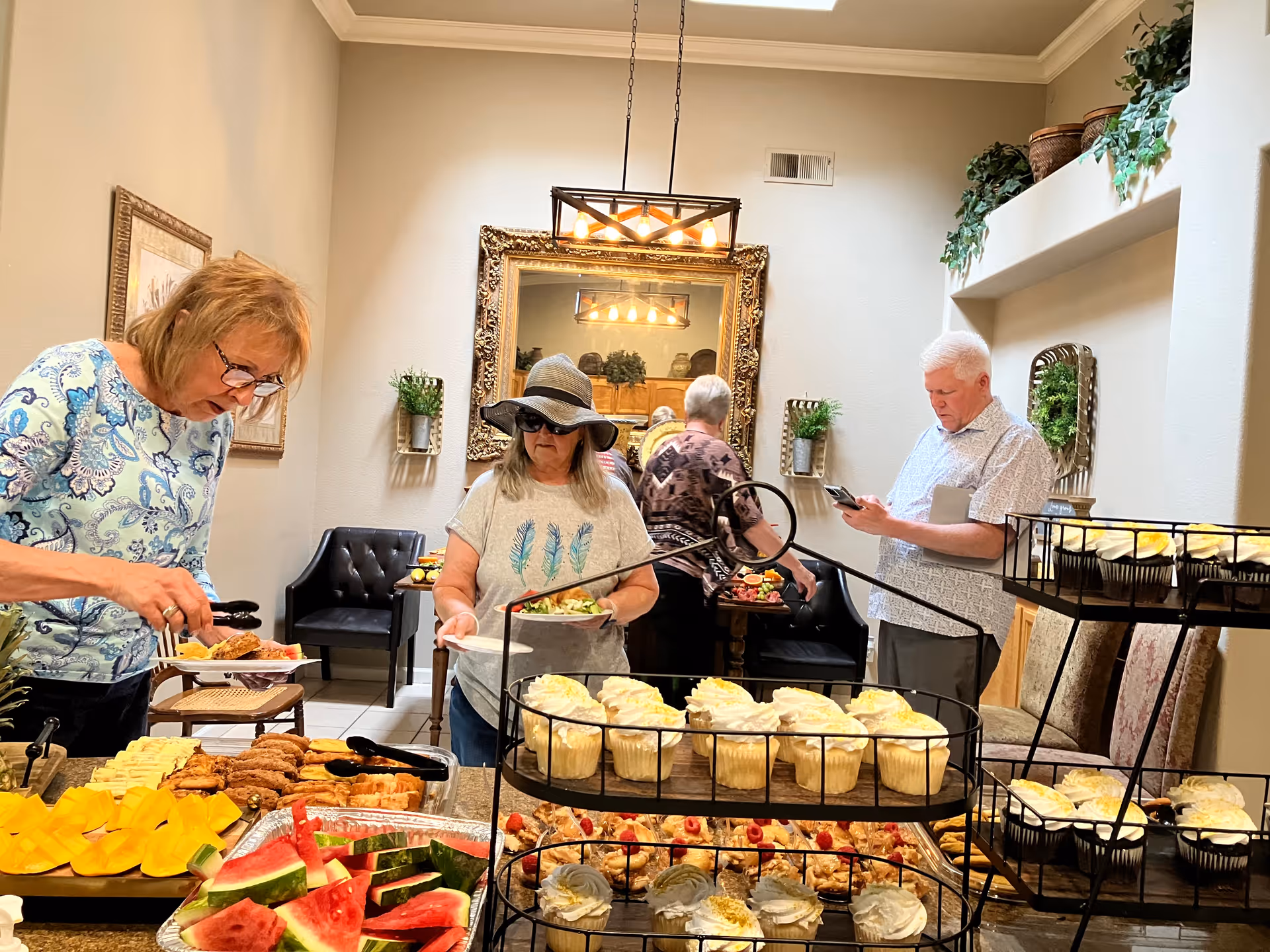 Four elderly people serving themselves food from a buffet table in a dining room. The buffet includes cupcakes, watermelon slices, and various other dishes. The room has beige walls, a large ornate mirror, plants on shelves, and a modern light fixture hanging from the ceiling.