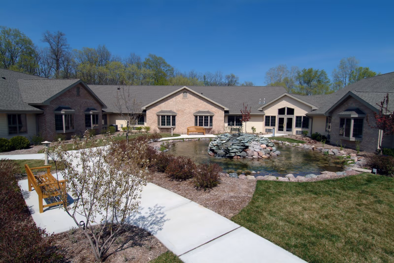 A landscaped courtyard with a central pond and rock fountain, surrounded by walkways, benches, shrubs, and a single-story brick building under a blue sky.