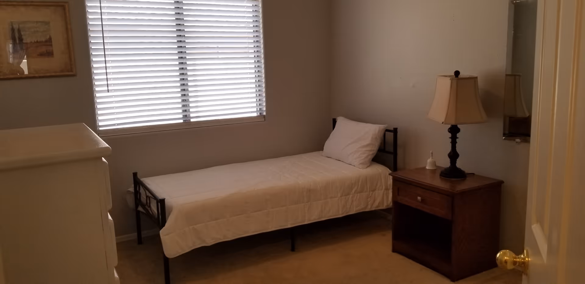 A small bedroom with a single bed covered with a white quilt and a white pillow. Next to the bed is a wooden nightstand with a lamp and a small bell. A window with closed blinds is on the wall behind the bed, and a framed picture hangs on the adjacent wall. A white dresser is partially visible on the left side of the image.