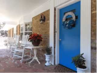 A covered porch area with a blue door decorated with a white and blue wreath. The porch has several white rocking chairs lined up against a brick wall, a small round table with a pot of red flowers, and two white planters with green plants. The floor is made of reddish stone tiles.