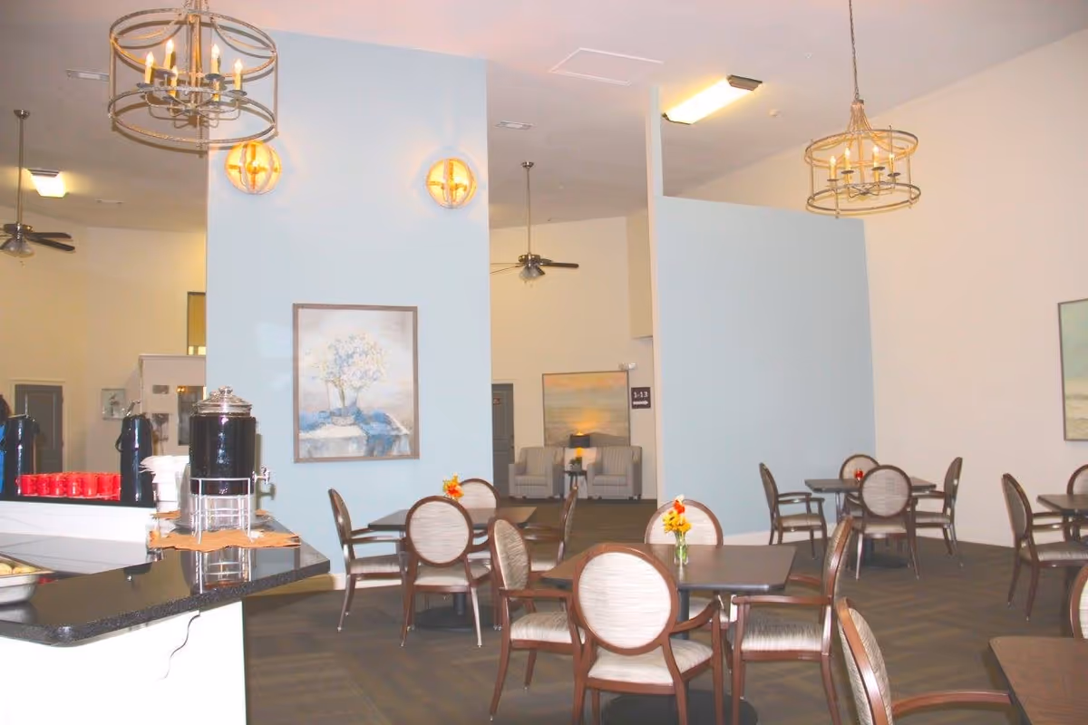 A bright dining area in a senior living facility with several round tables and cushioned chairs. Each table has a small vase with flowers. The room features light blue and white walls, modern chandeliers, wall sconces, and ceiling fans. There are framed paintings on the walls and a counter with beverage dispensers and cups.