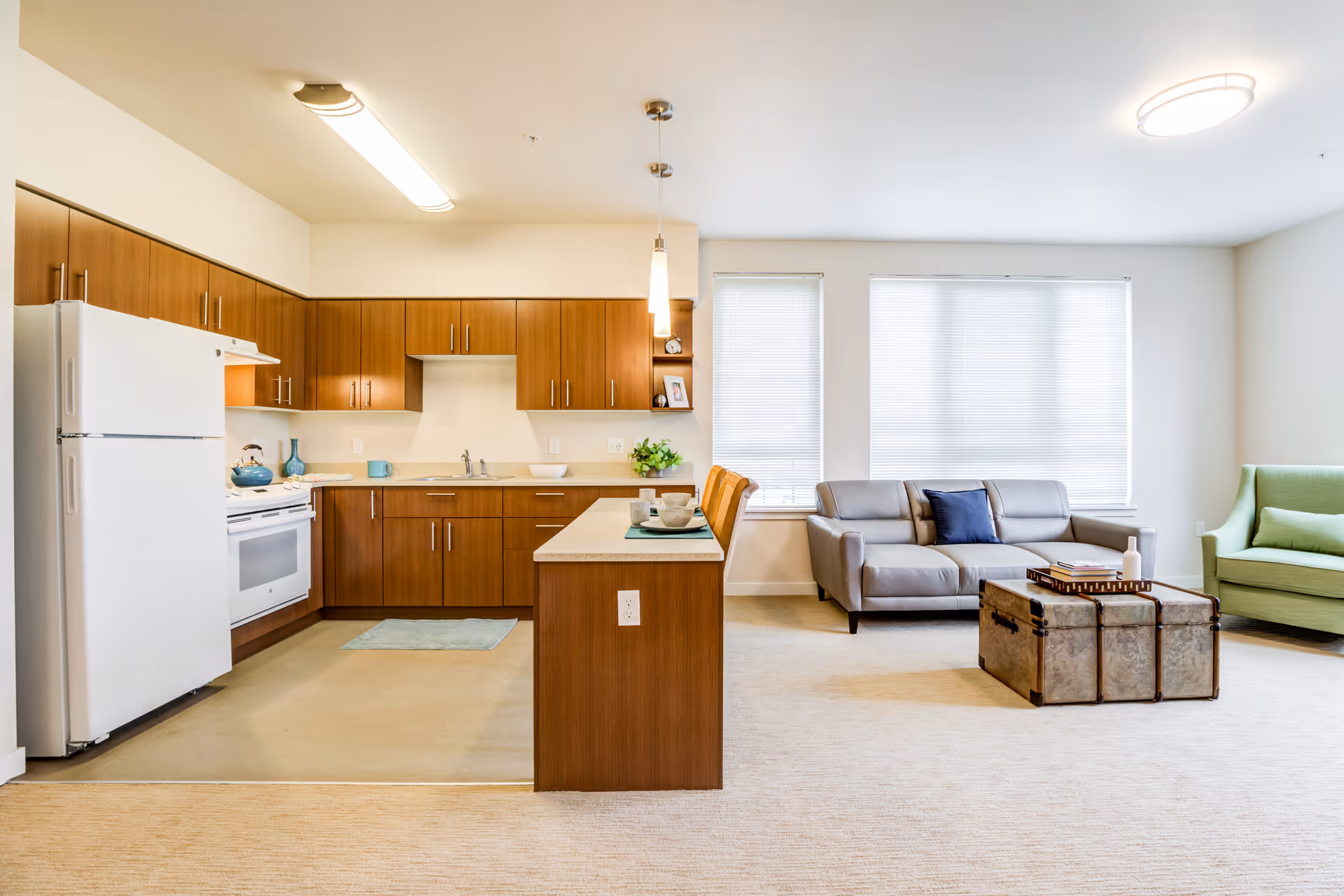 Open-plan interior showing a kitchen with wood cabinets and island on the left and a living area with a gray sofa, green armchair, and trunk coffee table on the right.