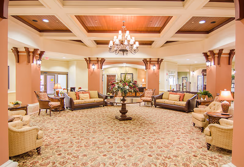 Spacious and elegantly decorated living room area with patterned carpet, multiple sofas and armchairs arranged around a central round table with a floral arrangement. The room features warm lighting, a chandelier hanging from a coffered ceiling, and decorative columns with sconces.
