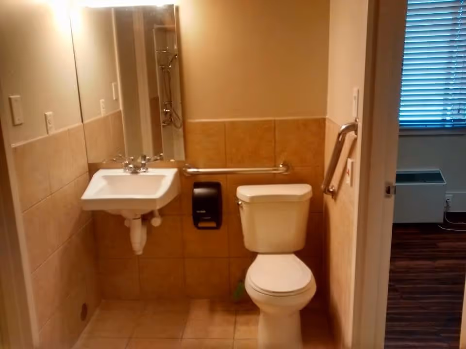A small bathroom with beige tiled walls and floor, featuring a white toilet and a wall-mounted white sink with a mirror above it. There are metal grab bars next to the toilet and sink for accessibility. A black paper towel dispenser is mounted on the wall between the sink and toilet. The bathroom door is open, showing a glimpse of a room with wooden flooring and a window with closed blinds.