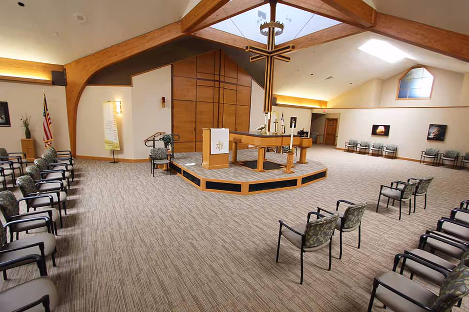 Interior view of a chapel or worship room with rows of chairs arranged facing a wooden altar with religious symbols. The room has a high ceiling with wooden beams, soft lighting, and religious decorations including a cross hanging above the altar and an American flag in the corner.