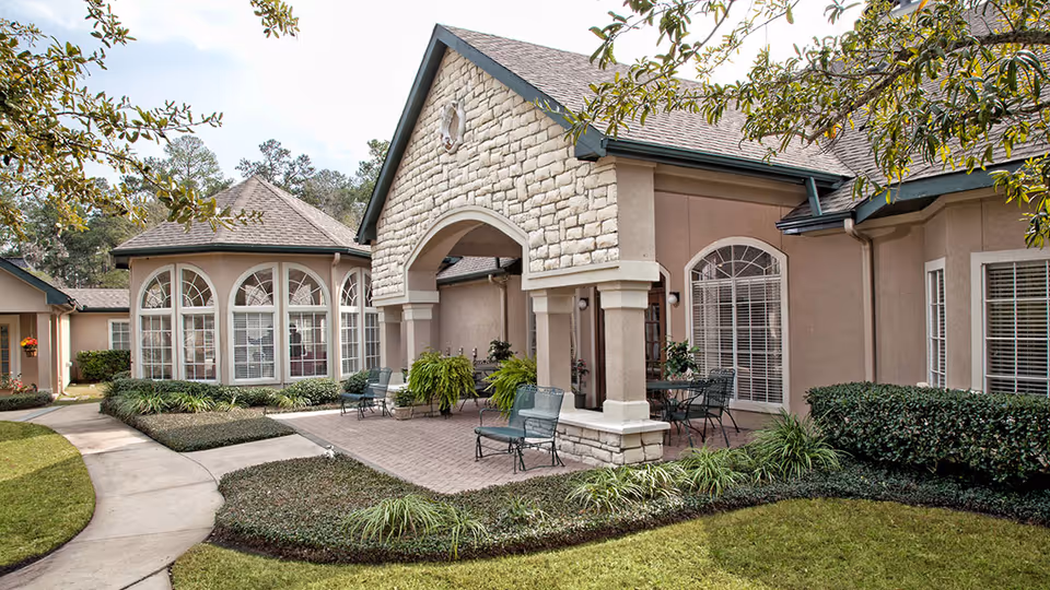 Outdoor patio area of a senior living facility with stone and stucco exterior walls, arched windows, and a covered seating area with metal chairs and tables surrounded by landscaped greenery and a curved concrete walkway.
